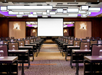 A modern conference room with rows of tables and chairs facing a large projector screen. The ceiling features geometric light fixtures and the space is illuminated with purple accent lighting.