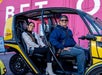 A man and woman smiling and sitting on in front of the other in a yellow door-less GoCar with a bright pink wall behind them.