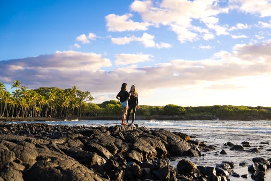 Two people stand on black volcanic rocks near the shoreline with palm trees and a partly cloudy sky in the background.