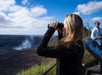 A woman uses binoculars to observe a volcanic crater emitting smoke, while another person stands nearby under a blue sky with clouds.