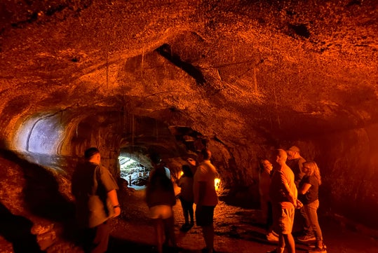 A group of people stands inside a dimly lit lava tube cave with rough, orange-lit walls and a bright opening visible in the distance.