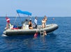Five people are on and around a small inflatable boat in the ocean, some wearing snorkeling gear, with clear blue water and sky in the background.