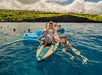 Several people enjoy paddleboarding and swimming in clear blue water near a rocky shore on a sunny day, with children and adults smiling at the camera.