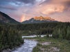 A river flows through a forest with a mountain backdrop under a colorful sky. A faint rainbow appears in the sky near the mountain.