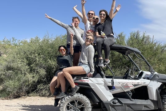 A group of young women sitting on the back of a white ATV smiling with their arms in the air and the desert behind them on a sunny day.