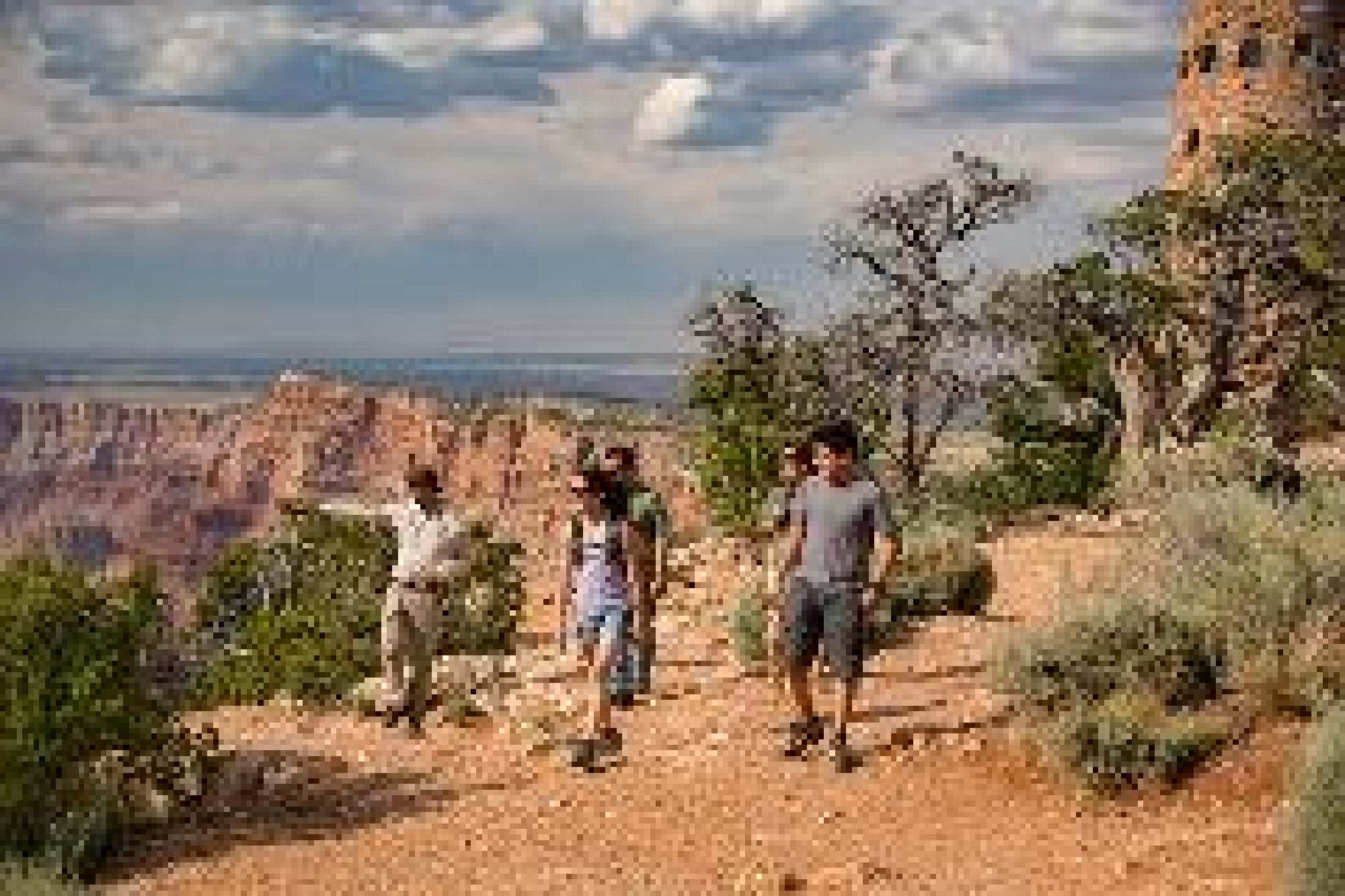 Four people walk along a trail with canyon views and sparse vegetation under a partly cloudy sky.