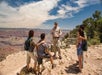 A group of five people stands on a rocky ledge overlooking a canyon, with one person speaking as the others listen. Trees and clouds are visible in the background.