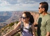 A young girl and an adult man wearing sunglasses stand at a scenic overlook, gazing at the Grand Canyon under a partly cloudy sky.