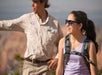 A young woman wearing sunglasses and a backpack smiles outdoors while a person in a park ranger uniform gestures in the background.