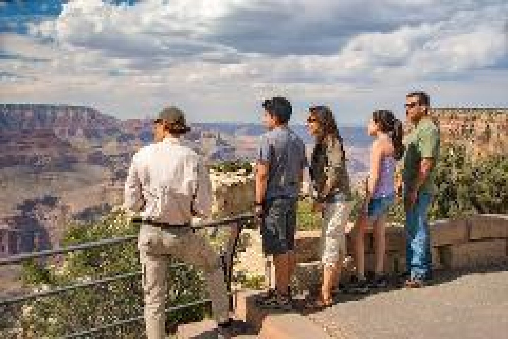 Five people stand at a viewpoint, looking out over the Grand Canyon under a partly cloudy sky.