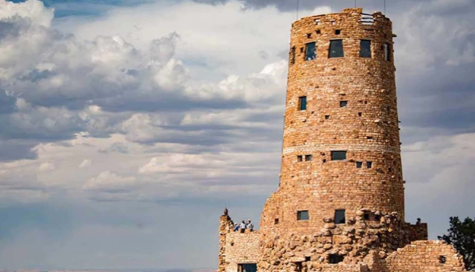 A cylindrical stone watchtower with small windows stands against a cloudy sky, with several people visible at its base.