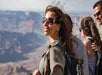 A group of people wearing backpacks and sunglasses stand at a scenic overlook with a canyon in the background.