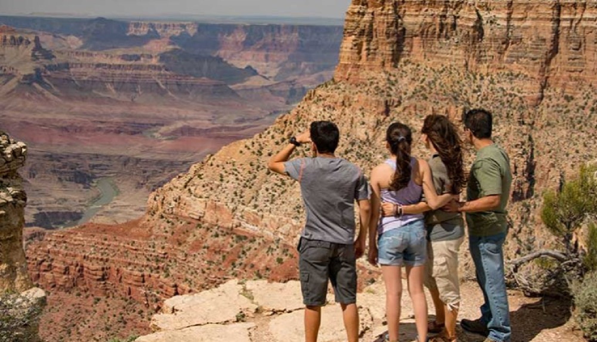 Four people stand at the edge of a rocky cliff overlooking the expansive Grand Canyon, with layered rock formations and a river visible below.
