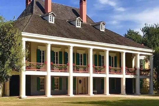 A large historic plantation-style house with white columns, a dark roof, green shutters, and multiple chimneys, set amidst greenery.