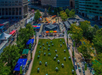 Aerial view of a city square with a green lawn, tables, and chairs. Colorful umbrellas surround a central fountain. Buildings and trees are in the background.