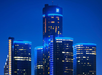 Night view of illuminated skyscrapers, with a prominent tower displaying the GM logo against a clear blue sky.