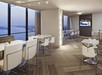 Modern lounge area with white chairs and tables, featuring a large window with a view of the water and a TV displaying a sports game.