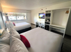 Hotel room with two neatly made beds, a wall-mounted TV, a desk, and storage area with a microwave and mini-fridge. Large window on the left side. Modern decor with wood flooring.