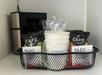 Coffee maker and a wire basket containing coffee packets, Tazo tea bags, and a stack of white cups wrapped in plastic, placed on a shelf.