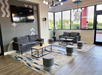 Modern waiting area with two gray sofas, two wooden coffee tables, and round gray stools on colorful rugs. Large TV mounted on wall; large windows let in natural light.