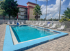 Outdoor rectangular swimming pool surrounded by white lounge chairs, with a red and beige building in the background under a partly cloudy sky.