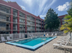 Outdoor swimming pool surrounded by lounge chairs and a multi-story red building with balcony railings, set against a clear blue sky with scattered clouds.