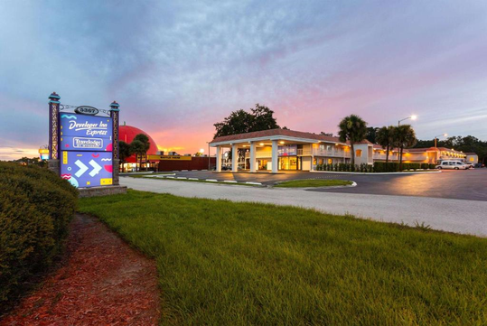A white building with a parking lot and palm trees at dusk. The foreground has grass and shrubs, and a sign reads "Developer Inn Express." A colorful sky is visible in the background.