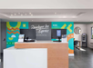 Reception area of a hotel with a modern design, featuring two computers on the counter, colorful wall graphics, and an ATM in the corner.