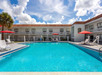 Outdoor hotel pool with clear blue water, surrounded by white lounge chairs, red umbrellas, and two-story building under a partly cloudy sky.