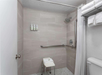 Accessible shower with beige tiles, featuring a showerhead, grab bars, toiletries on the wall, a white curtain, and a small stool with folded towels.