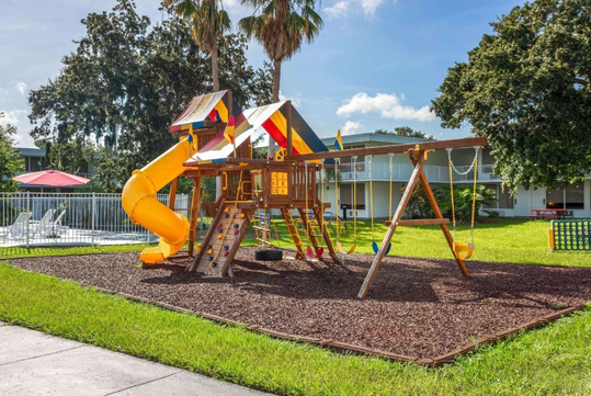 A colorful playground set with a slide, swings, and climbing structures sits on a mulch surface, surrounded by green grass and backed by palm trees and buildings.