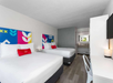 Hotel room with two double beds, a desk with chair, and a wall-mounted TV. The beds have colorful headboards and red pillows. A sink area is visible in the background.