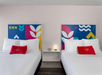 Two neatly made beds with white linens and red pillows in a hotel room. Colorful geometric artwork hangs above each bed, and a small white nightstand with a round lamp is between them.