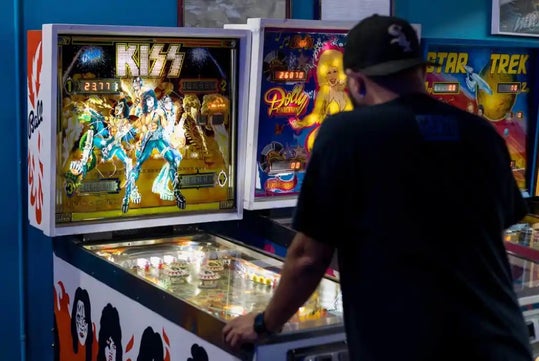 A person plays a Kiss-themed pinball machine in an arcade, with other pinball machines visible in the background.