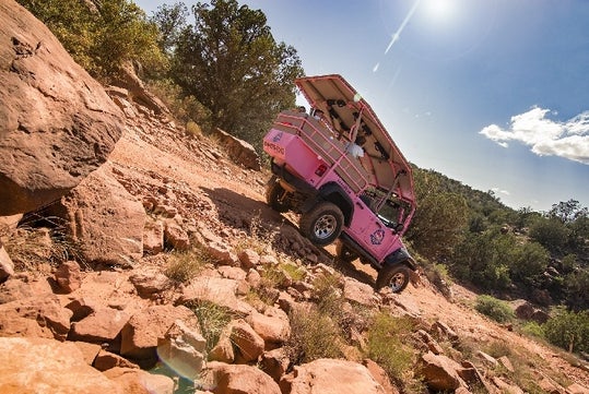 A pink off-road tour vehicle drives up a steep, rocky slope in a desert landscape under a bright, sunny sky.