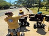 A group of people wearing helmets ride Segways on a sidewalk while one person points ahead, leading the group.