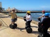 A group of people wearing helmets ride Segways along a seaside path, while one person appears to lead or instruct the group.