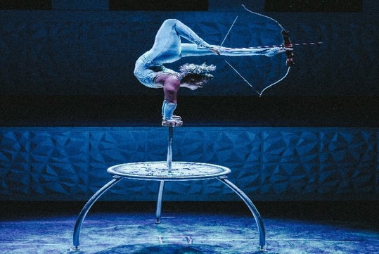 A contortionist balances on one hand atop a raised platform while holding a bow and aiming an arrow with their feet.