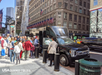 A group of people wait to board a black USA Guided Tours bus parked on a busy city street lined with American flags and tall buildings.