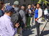 A woman speaks to a group of people standing in line on a sidewalk in daytime; some people are looking at their phones.