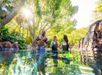 Three people stand in a clear, tropical pool surrounded by lush greenery, holding drinks and talking under bright sunlight.