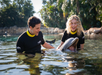 Two people in wetsuits interact with a dolphin in shallow water at a marine park, with trees and rocks visible in the background.
