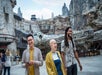 Three people walk and smile in front of the Millennium Falcon at a Star Wars-themed amusement park, with others and rocky structures in the background.