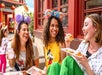 Three women wearing Mickey Mouse ear headbands sit outside, smiling and eating food from plates. A child in the background holds a drink. The setting appears to be a theme park or festive location.