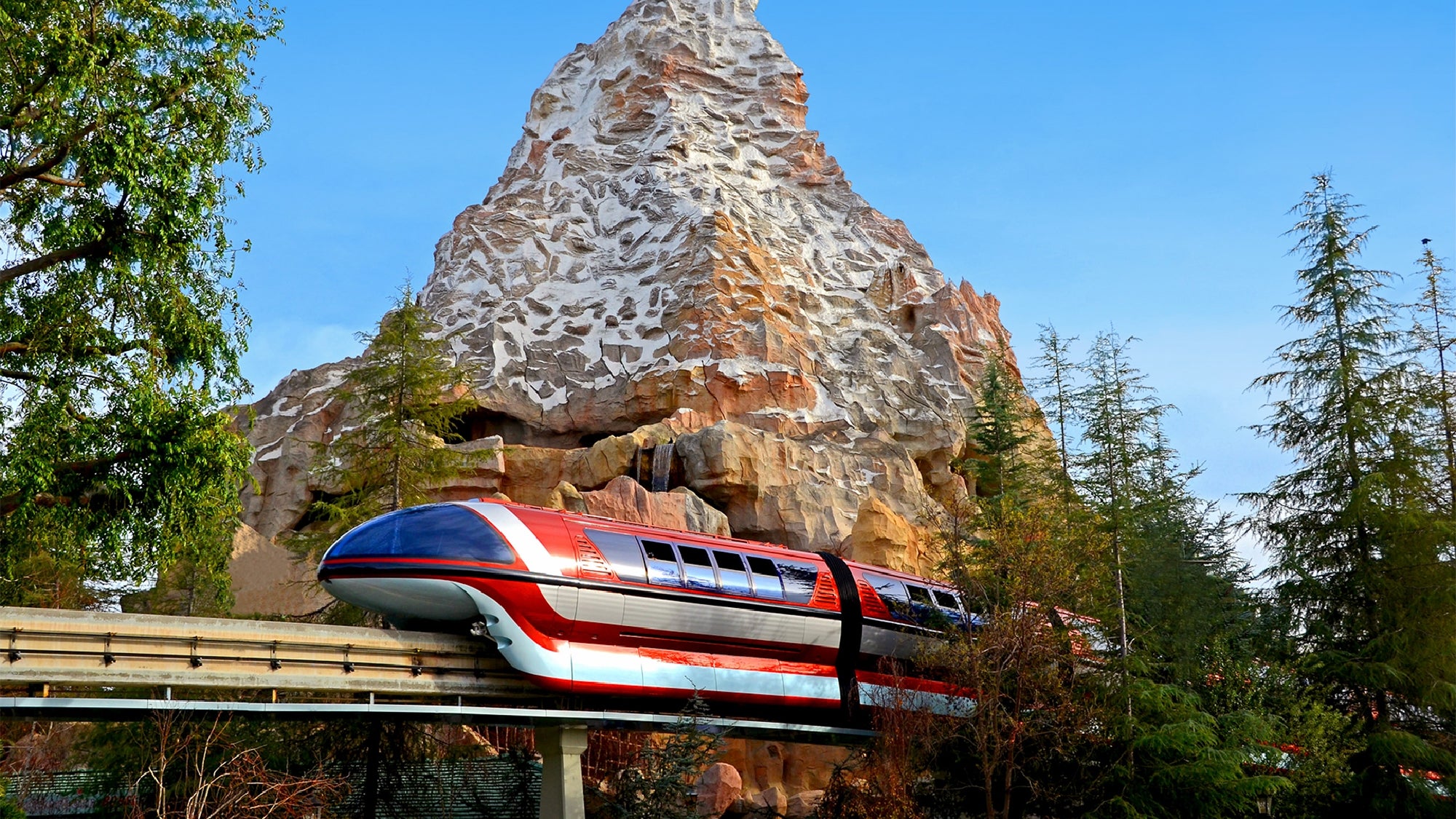 A red, white, and blue monorail travels on an elevated track in front of a rocky, snow-capped mountain surrounded by trees.