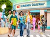 Four people walk and smile outside the Mickey & Minnie's Runaway Railway attraction at a theme park, with other visitors and colorful buildings visible in the background.