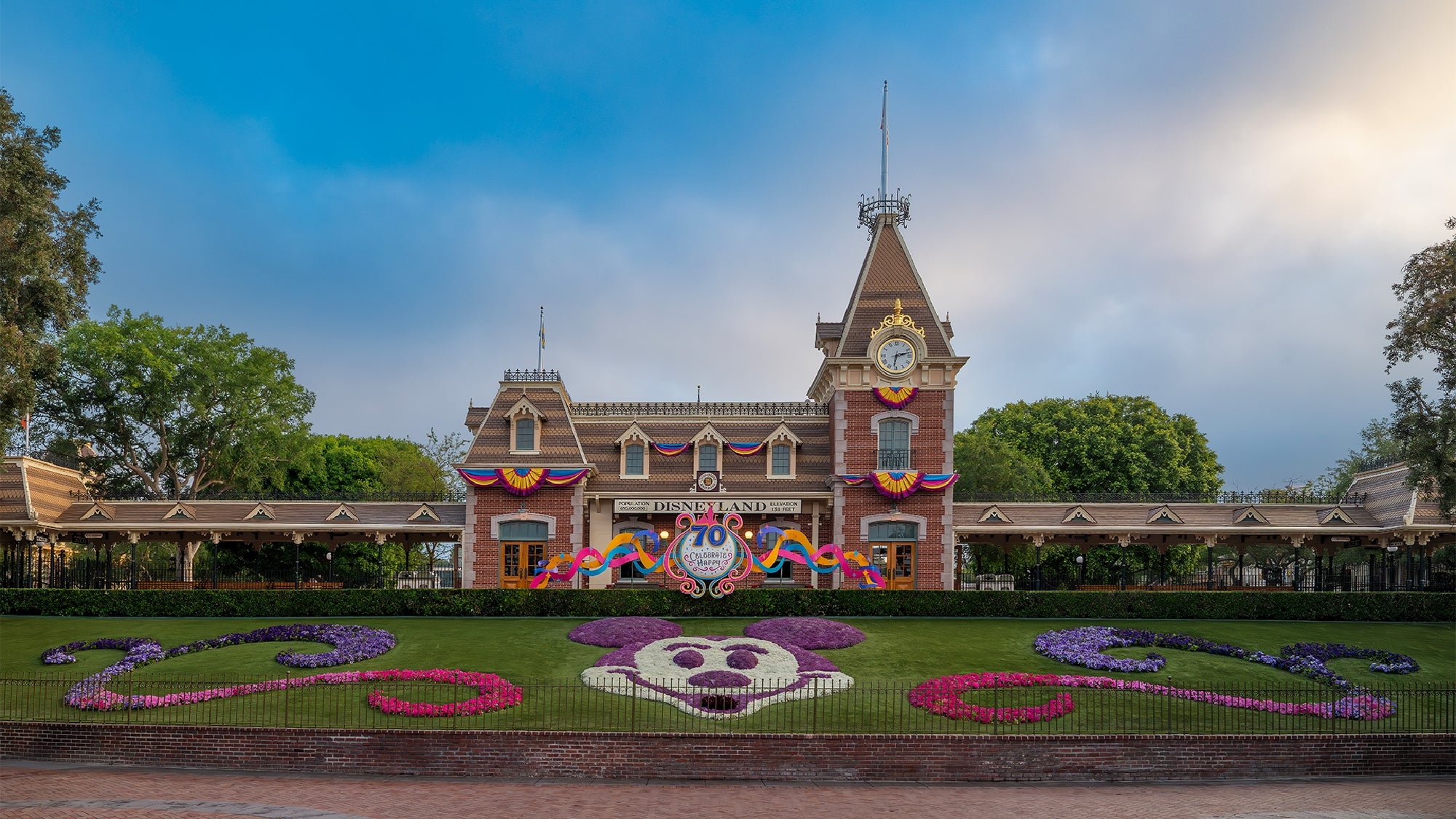 Disneyland entrance with a floral Mickey Mouse design on the lawn, a decorated train station building, and a 70th anniversary banner.