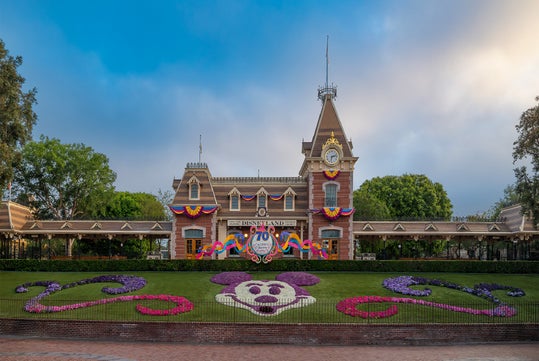 Disneyland entrance with a floral Mickey Mouse design on the lawn, a decorated train station building, and a 70th anniversary banner.