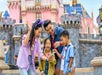 A family of four smiles and looks at a smartphone in front of a pink castle at an amusement park.