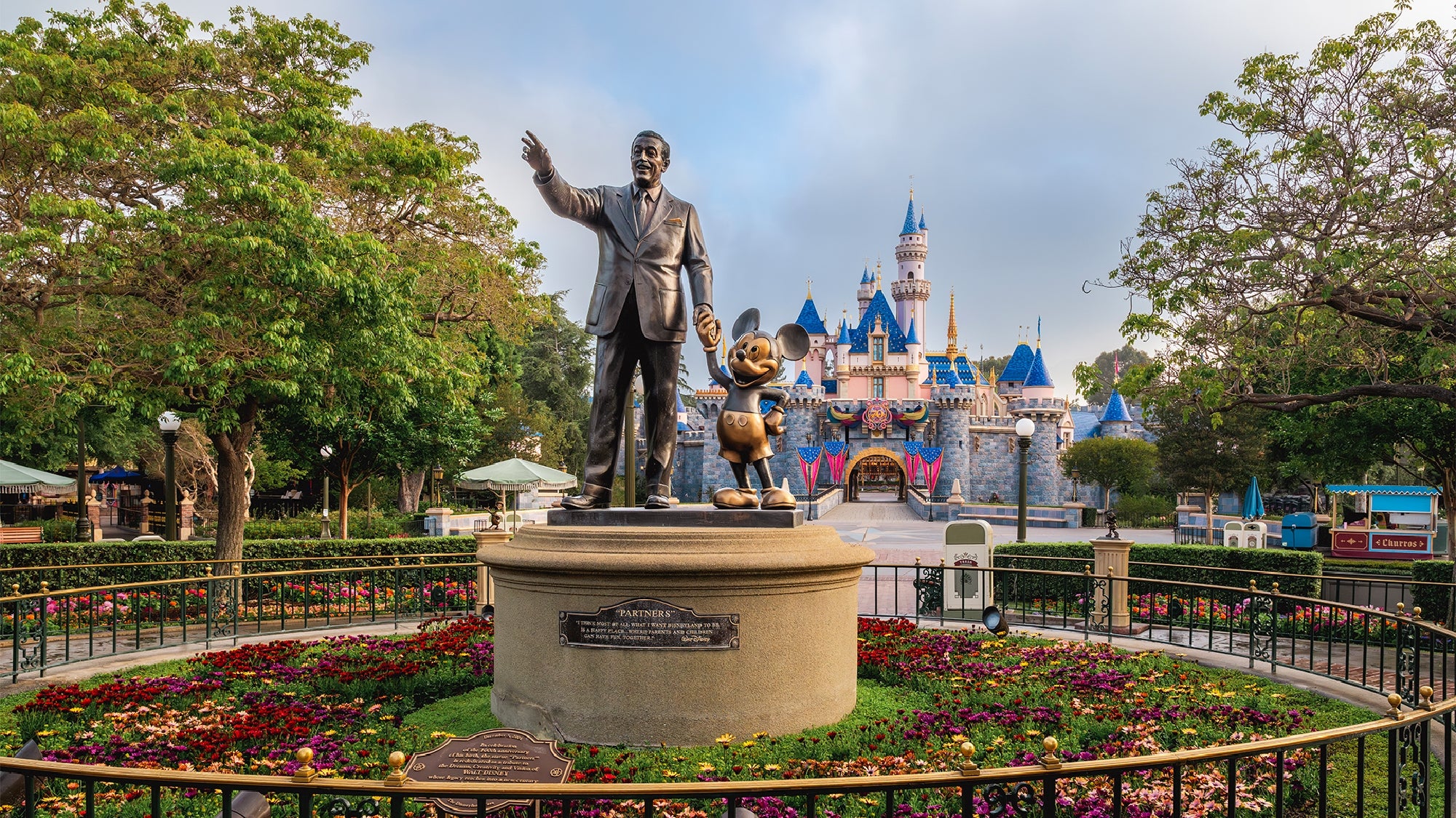 Bronze statue of a man and Mickey Mouse stands in front of a castle, surrounded by flowers and trees, at the entrance to Disneyland.